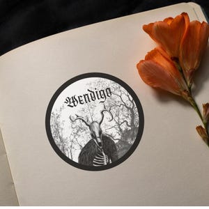 May include: A circular sticker with the word "Wendigo" in gothic lettering. The design features a skeletal figure with antlers and a fur cloak, set against a forest scene. An orange flower is placed on the page.
