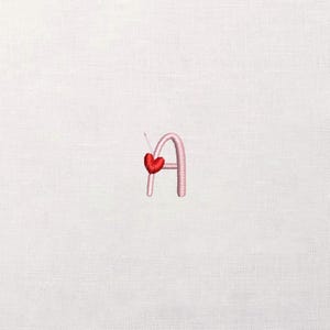 May include: Embroidered letter "A" in light pink, with a red heart attached. The heart is stitched with red thread. The background is a plain white fabric.