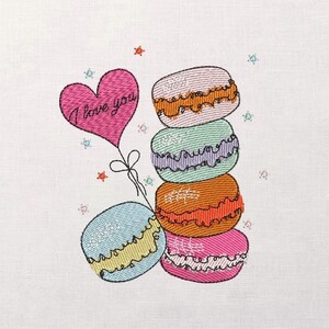Puede incluir: Un diseño bordado con una pila de macarons coloridos y un globo rosa en forma de corazón. El globo tiene el texto "I love you". Los macarons son de color rosa, naranja, verde y azul, con costuras decorativas.