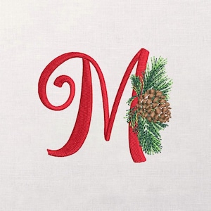 May include: Embroidered red letter "M" with a decorative pinecone and evergreen branch design. The letter is in a cursive font, and the pinecone is brown with green needles. The background is white.