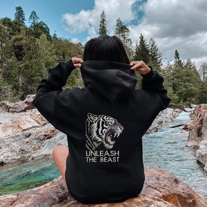 May include: Black hoodie with a white tiger graphic and the text "UNLEASH THE BEAST". The hoodie is made of a soft material and has a hood. The person is sitting on a rock near a river.