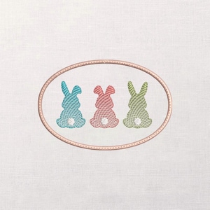 May include: An embroidered design featuring three colorful bunny silhouettes in blue, pink, and green, each with a white tail. The bunnies are enclosed within a light brown oval frame, set against a white fabric background.