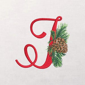 Peut inclure: Lettre "J" rouge brodée avec un motif festif. La lettre est ornée d'aiguilles de pin vertes et d'une pomme de pin marron, créant un thème de vacances ou de saison. Le motif est posé sur un fond blanc.