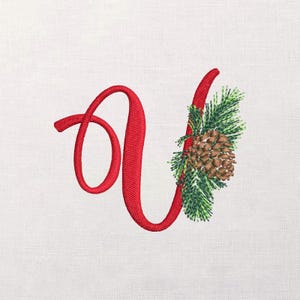 Może przedstawiać: Haftowana czerwona litera "A" z dekoracyjną szyszką i gałązką iglastą. Projekt znajduje się na białym tle, idealny do świątecznych rękodzieł i dekoracji.