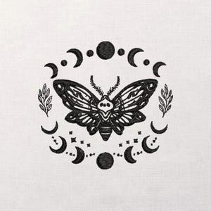 May include: Black and white embroidered design featuring a moth with a skull-like pattern on its wings, surrounded by crescent moons, stars, and circles. Two sprigs of leaves are on either side of the moth. The design is on a white fabric.