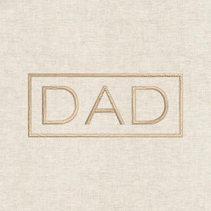 May include: Cream-colored fabric with the word "DAD" embroidered in a gold color. The word is inside a gold rectangular frame. The embroidery has a textured appearance.