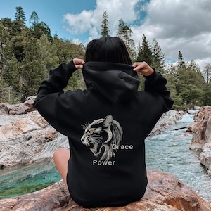 Pode incluir: Moletom preto com um design detalhado de pantera nas costas, acompanhado das palavras "Grace in Power" em branco. A pessoa está sentada em uma rocha perto de um rio, com árvores e um céu nublado.