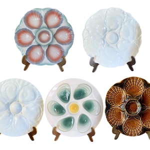 Pode incluir: Cinco pratos decorativos para servir ostras em várias cores e designs. Os pratos são redondos com vários compartimentos para ostras e um poço central. Cada prato é suportado por um pequeno suporte castanho. Os pratos apresentam padrões de conchas e florais.