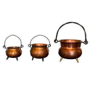 May include: A set of three graduated copper pots with a hammered texture. Each pot features a rounded body, three small legs, and a dark metal swing handle attached to the sides. The pots vary in size from smallest to largest, displaying a metallic finish.