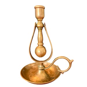 May include: A vintage brass candlestick with a unique design. The candle holder is shaped like a teardrop with a round ball at the top. The base is a circular tray with a handle.