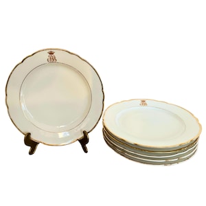 May include: A collection of white porcelain plates with a gold rim and a scalloped edge. Each plate has a gold emblem and a gold inner ring. One plate is on a black stand, the others are stacked.
