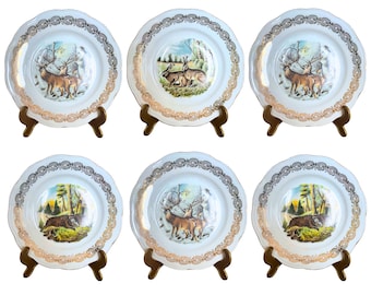 May include: Set of six white ceramic dinner plates with gold trim and woodland animal illustrations. Each plate features a different animal, including deer, rabbits, and a boar.