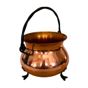 May include: A polished copper cauldron with a rounded body, featuring a dark metal swing handle attached to the sides and three dark metal feet at the base. The surface of the copper pot is smooth and reflective, showing a warm metallic tone.