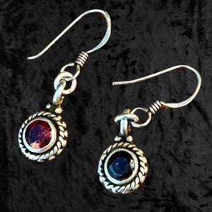 May include: A pair of silver earrings with a round, dark red gemstone on one earring and a round, dark blue gemstone on the other earring. The earrings have a silver hook closure.