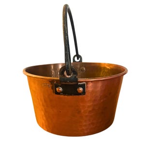 May include: A hammered copper pot with a black iron handle. The pot has a rounded bottom and a wide opening.