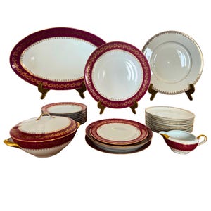 May include: A collection of fine china with a burgundy and gold floral design. The set includes an oval platter, dinner plates, soup tureen, saucers, and a gravy boat. The plates are displayed on small wooden stands.