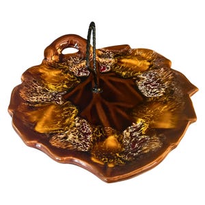 Puede incluir: Una fuente de cerámica marrón con forma de hoja y asa de metal. La fuente tiene un acabado brillante con una mezcla de esmalte marrón, dorado y blanco. El asa es de metal oscuro y está unida al centro de la fuente.