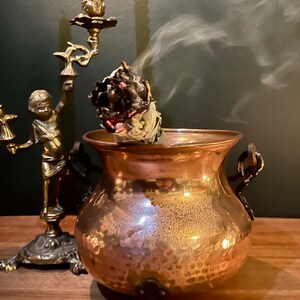 Può includere: Un calderone di rame con tre gambe e un manico su ogni lato. Un bastoncino di salvia bianca fumante sta bruciando all'interno del calderone, con fumo che sale.