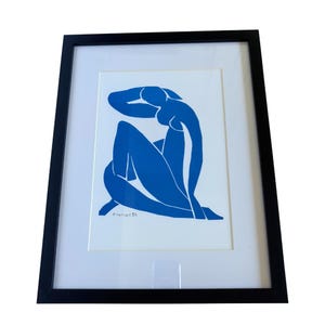 Op de afbeelding: Een ingelijste prent met een blauw silhouet van een zittende figuur. Het kunstwerk staat tegen een witte achtergrond, omlijst met een zwarte rand. De prent is gesigneerd en gedateerd.