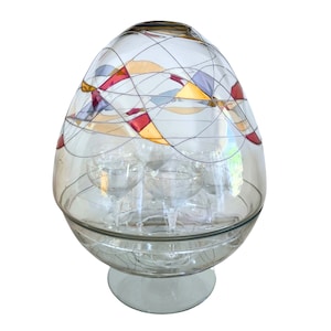 May include: A clear glass dome with a geometric pattern of red, gold, and blue lines. The dome sits on a clear glass base and holds six clear glass cups.