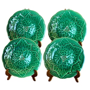 May include: Four green ceramic plates with a leaf design. Each plate has a central leaf pattern surrounded by a textured border. The plates are displayed on gold-colored stands, showcasing their intricate details and vibrant color.