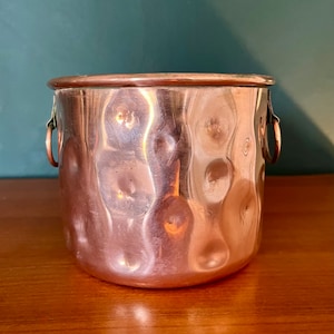 Peut inclure: Un pot en cuivre martelé avec deux petites poignées en boucle. Le pot a une surface brillante et réfléchissante.