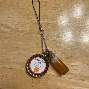 May include: A silver charm with a bottle filled with a light brown liquid and a bottle cap with the text "Pumpkin Spice Latte" and a cartoon image of a pumpkin spice latte.