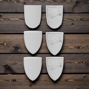 Op de afbeelding: Zes witte schildvormige objecten, drie effen en drie met een ontwerp, gerangschikt op een donker oppervlak met houtnerf. De schilden zijn symmetrisch met een afgeronde bovenkant en een puntige onderkant. De ontworpen schilden hebben een rechthoekig element met lijnen.