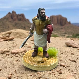 May include: A miniature figurine of a warrior, clad in gold armor and a white skirt, holding a sword and a red cloak. The figure stands on a circular base with a sandy texture, with a desert landscape in the background.