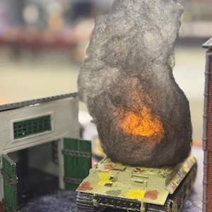 May include: A model tank with camouflage paint is positioned in front of a miniature building. A large, dark gray cloud-like structure with an orange glow sits atop the tank, suggesting an explosion. The scene is set on a cobblestone surface.