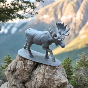 May include: A grey moose figurine standing on a grey rock with a mountain background.