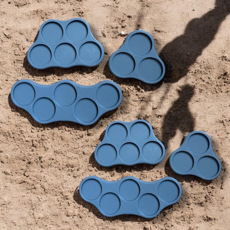 May include: Six blue plastic trays with multiple circular indentations, designed for organizing and storing small items. The trays are arranged on a sandy surface.