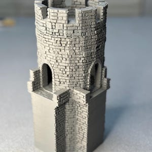 May include: A gray miniature castle tower with a detailed brickwork design. The tower features arched windows and a crenelated top, showcasing a medieval architectural style. The model is a solid, detailed structure.