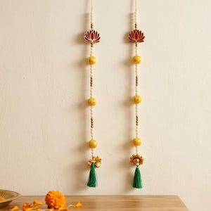 Puede incluir: Dos colgantes de pared verticales con una pieza superior en forma de loto rojo, cuentas amarillas y blancas alternadas y una borla verde en la parte inferior. Los cordones decorativos cuelgan simétricamente contra una pared lisa.