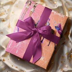 Può includere: Una confezione regalo avvolta in carta rosa con illustrazioni di figure. La scatola è legata con un ampio nastro di raso viola scuro e un fiocco. Lo sfondo è un tessuto color crema con accenti di stelle dorate.
