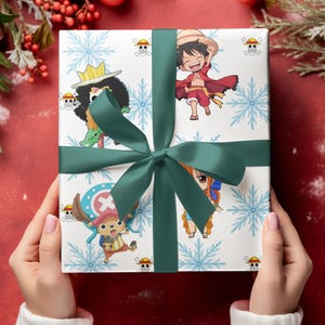 Pu&ograve; includere: Un regalo confezionato con un nastro e un fiocco verdi, con personaggi della serie anime One Piece. La carta da regalo &egrave; bianca con fiocchi di neve blu e illustrazioni di personaggi. Il regalo &egrave; tenuto su uno sfondo rosso con decorazioni festive.