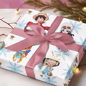 Pu&ograve; includere: Regalo confezionato con carta bianca decorata con personaggi dei cartoni animati e fiocchi di neve blu. Un nastro e un fiocco rosa polveroso adornano il regalo. I personaggi sono della serie anime One Piece.