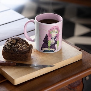 May include: A pink ceramic mug with a handle features a colorful anime character with pink hair and a green and purple outfit. The mug is filled with coffee and steam is rising from the top. A brown muffin is on a wooden cutting board next to the mug.