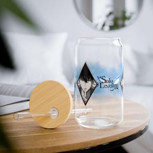 Può includere: Un bicchiere trasparente a forma di lattina con coperchio e cannuccia in bambù. Il bicchiere presenta una grafica in bianco e nero di una persona con la scritta "Solo Leveling" in blu. Il coperchio e il tavolo sono in legno.