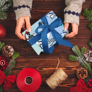 Può includere: Un regalo avvolto con un nastro blu è tenuto tra le mani, circondato da decorazioni natalizie su una superficie di legno. Il regalo ha una carta azzurra con un design grafico e la scritta "So Feeling". Ornamenti rossi, pigne e verde completano la scena festosa.