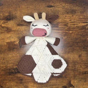May include: A crocheted cow-shaped baby blanket with brown and white spots. The blanket has a pink nose and closed eyes.