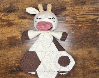 Op de afbeelding: Een gehaakt babydekentje in de vorm van een koe met bruine en witte vlekken. Het dekentje heeft een roze neus en gesloten ogen.