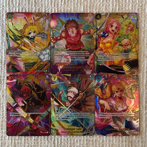 Puede incluir: Seis cartas coleccionables de One Piece dispuestas en dos filas. Las cartas presentan ilustraciones coloridas de personajes como Carrot, Monkey D. Luffy, Rebecca, Shanks, Trafalgar Law y Charlotte Pudding, cada una con texto impreso y estadísticas.