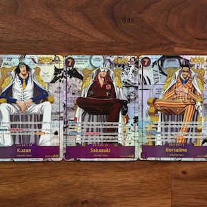 Puede incluir: Tres cartas coleccionables con personajes de manga. Cada carta muestra un personaje sentado, con texto e ilustraciones. Los personajes se llaman Kuzan, Sakazuki y Borsalino. Las cartas tienen un borde morado y están sobre madera.
