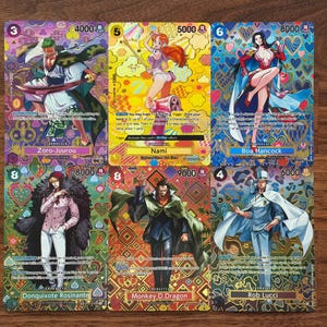 May include: Six colorful trading cards featuring anime characters. Each card displays a character illustration, character name, and numerical values. The cards have vibrant backgrounds with intricate patterns and are arranged on a wooden surface.