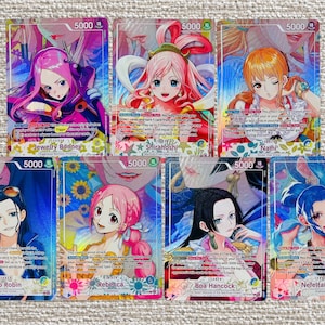 Puede incluir: Una colección de siete cartas coleccionables de One Piece con ilustraciones estilo anime de personajes femeninos, incluyendo a Jewelry Bonney, Shirahoshi, Nami, Nico Robin, Rebecca, Boa Hancock y Nefertari Vivi, cada una con un nivel de poder de 5000.