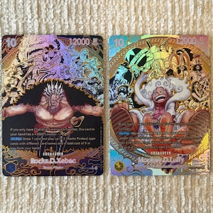 Può includere: Due carte collezionabili One Piece con dettagli in lamina olografica. La carta a sinistra raffigura Rocks D. Xebec con un costo di 10 e 12000 di potenza. La carta a destra mostra Monkey D. Luffy in forma Gear 5 con testo stampato.