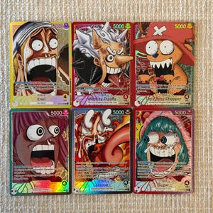 Puede incluir: Seis cartas coleccionables de One Piece dispuestas en dos filas. Las cartas muestran a los personajes Enel, Monkey D. Luffy, Tony Tony Chopper, Perona, Jozu y Sugar, cada uno con un nivel de poder de 5000, expresiones faciales y detalles holográficos.