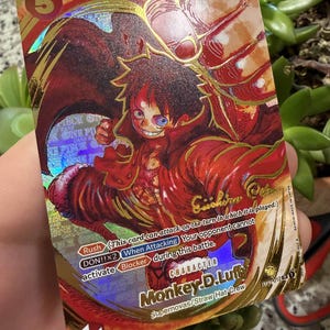 Puede incluir: Una tarjeta coleccionable con Monkey D. Luffy de los Supernovas/Tripulación del Sombrero de Paja. La tarjeta tiene un diseño de lámina dorada con una combinación de colores rojo y naranja. El texto de la tarjeta incluye "Rush", "DON!!x2" y "Blocker".
