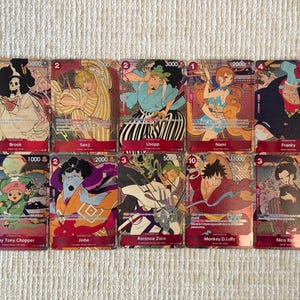 Puede incluir: Una colección de diez cartas coleccionables de One Piece dispuestas en dos filas, con personajes ilustrados que incluyen a Brook, Sanji, Usopp, Nami, Franky, Tony Tony Chopper, Jinbe, Roronoa Zoro, Monkey D. Luffy y Nico Robin con bordes rojos.